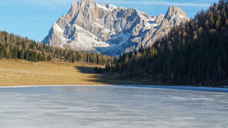 A frozen lake below a great pale Dolomite massif, larch and fir woods on either side