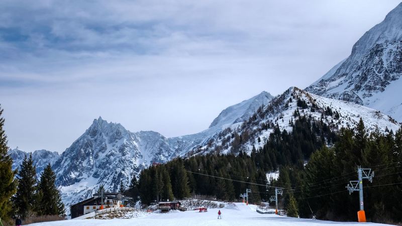 A wide groomed piste through the firs, a mountain restaurant and snowy peaks beyond