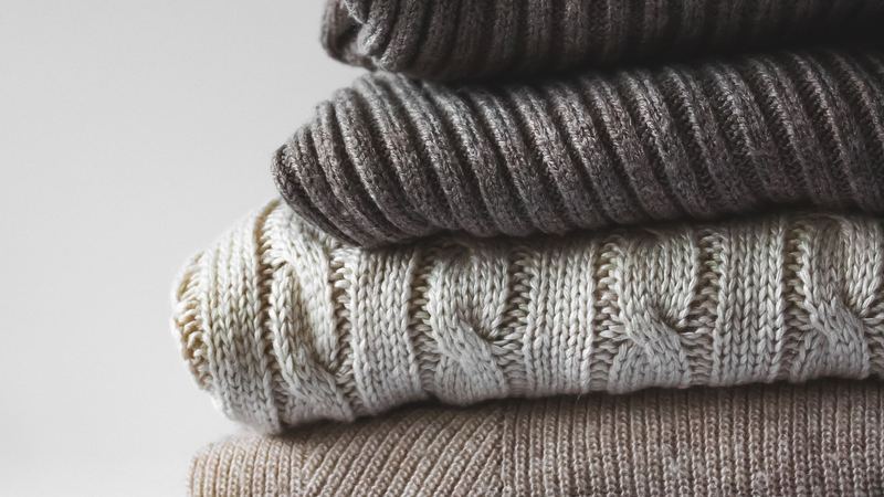 A stack of folded knitwear in grey, cream and camel, cable knit and rib