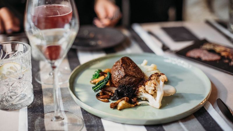 A fillet of beef with roasted cauliflower, wild mushrooms and kale on a pale plate, a glass of wine beside it