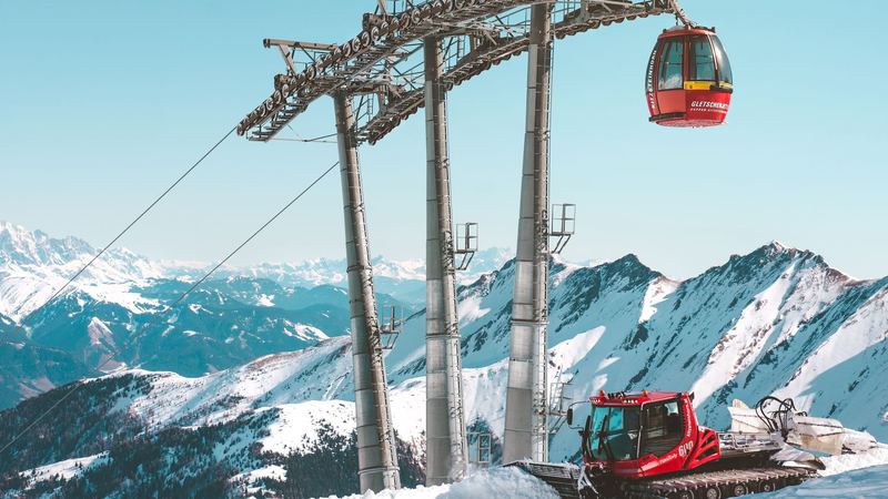 A red Gletscherjet gondola and a piste groomer high on the Kitzsteinhorn glacier above Kaprun, with snowy peaks beyond