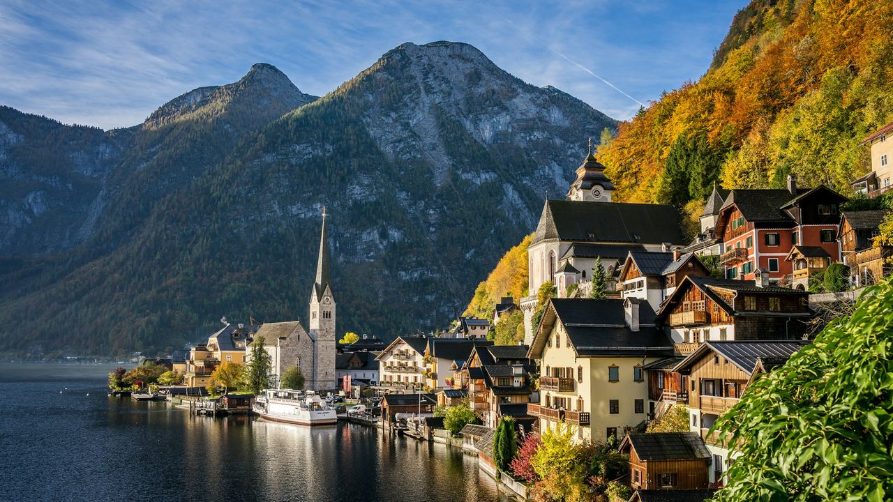 Hallstatt, its church spire and wooden houses on the still lake below wooded mountains in autumn