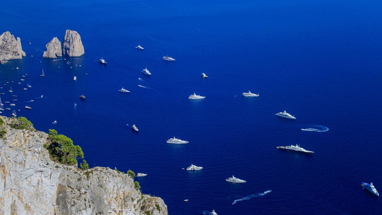 Superyachts at anchor in the deep blue sea off Capri, below the white cliffs and the Faraglioni rocks