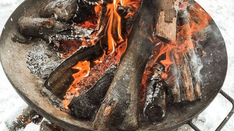 Logs blazing in an iron fire bowl on the snow, more firewood stacked beneath it