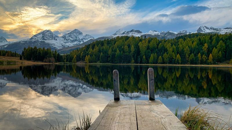 A wooden jetty on Lake Staz near St. Moritz, larch forest and snowy peaks reflected in the water