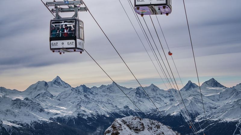 Two cable cars climbing above the snow, with the peaks of the Valais across the valley