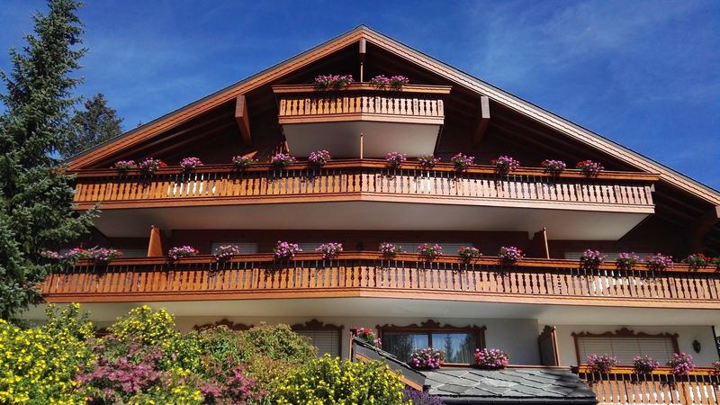 A wooden chalet with long balconies hung with pink geraniums, below a deep blue sky