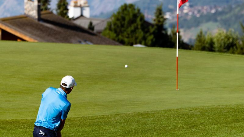 A golfer chipping onto the green towards the red flag, chalets and mountains beyond