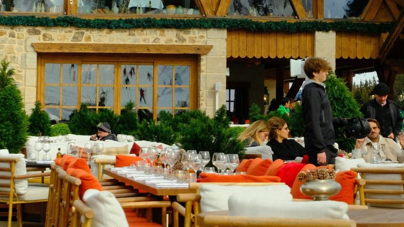 A mountain restaurant terrace laid with glasses and orange cushions in front of a timber chalet