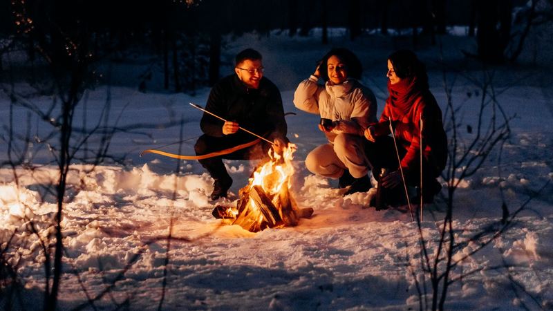 Three friends in winter coats gathered round a campfire in a snowy forest at night