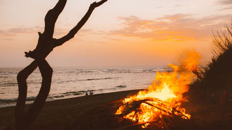 A bonfire blazing on a beach at sunset, the sea and a driftwood branch beyond