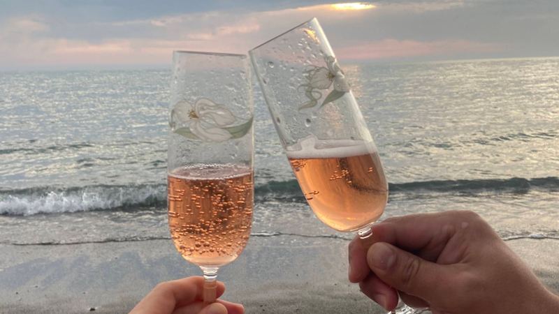 Two glasses of sparkling rosé raised on the beach at sunset, the sea behind