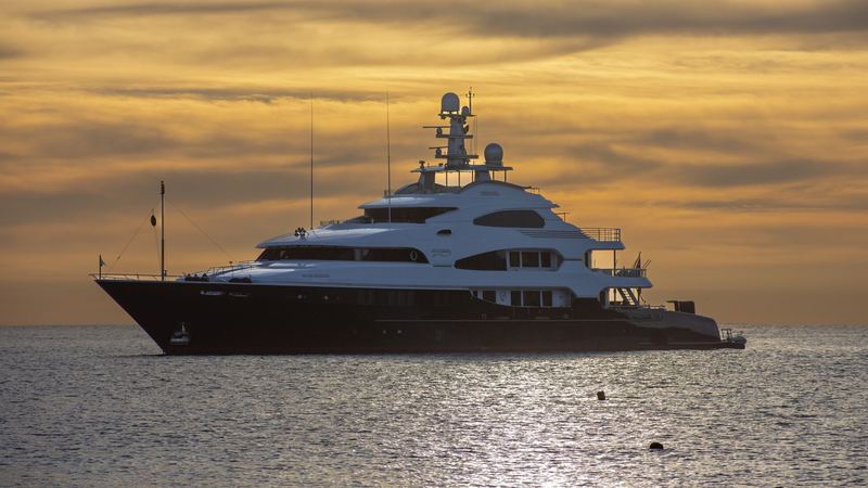 A superyacht at anchor on a silver sea under a golden sunset sky