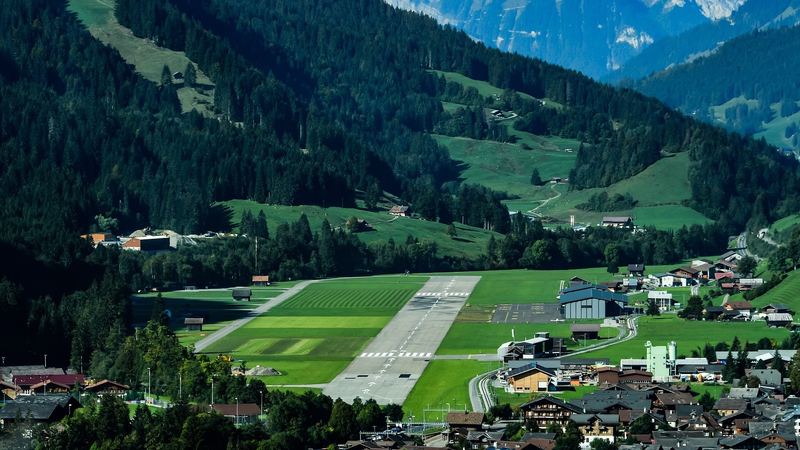The runway of Gstaad-Saanen airfield in a green valley between forested mountains, beside the chalets of Saanen