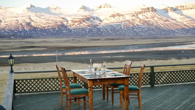 A wooden table laid with candles and glasses on a deck facing snow-covered mountains at dusk
