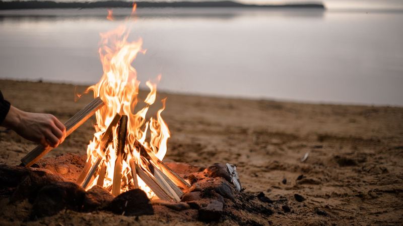 A hand adding wood to a fire on a sandy shore, still water beyond