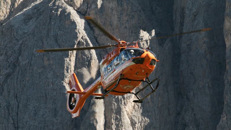 A red helicopter flying close to a sheer Dolomite rock wall