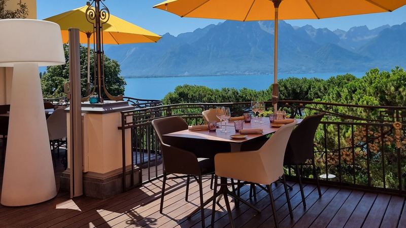 A terrace set for lunch under yellow parasols above a blue lake and the mountains