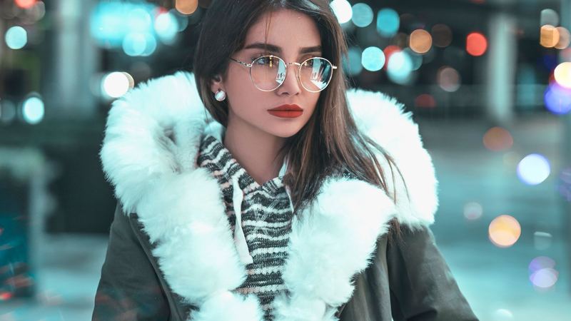 A woman in a winter coat with a white fur collar, the lights of the town blurred behind her at night