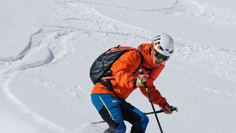 A skier in an orange jacket and helmet turning in deep powder off the piste