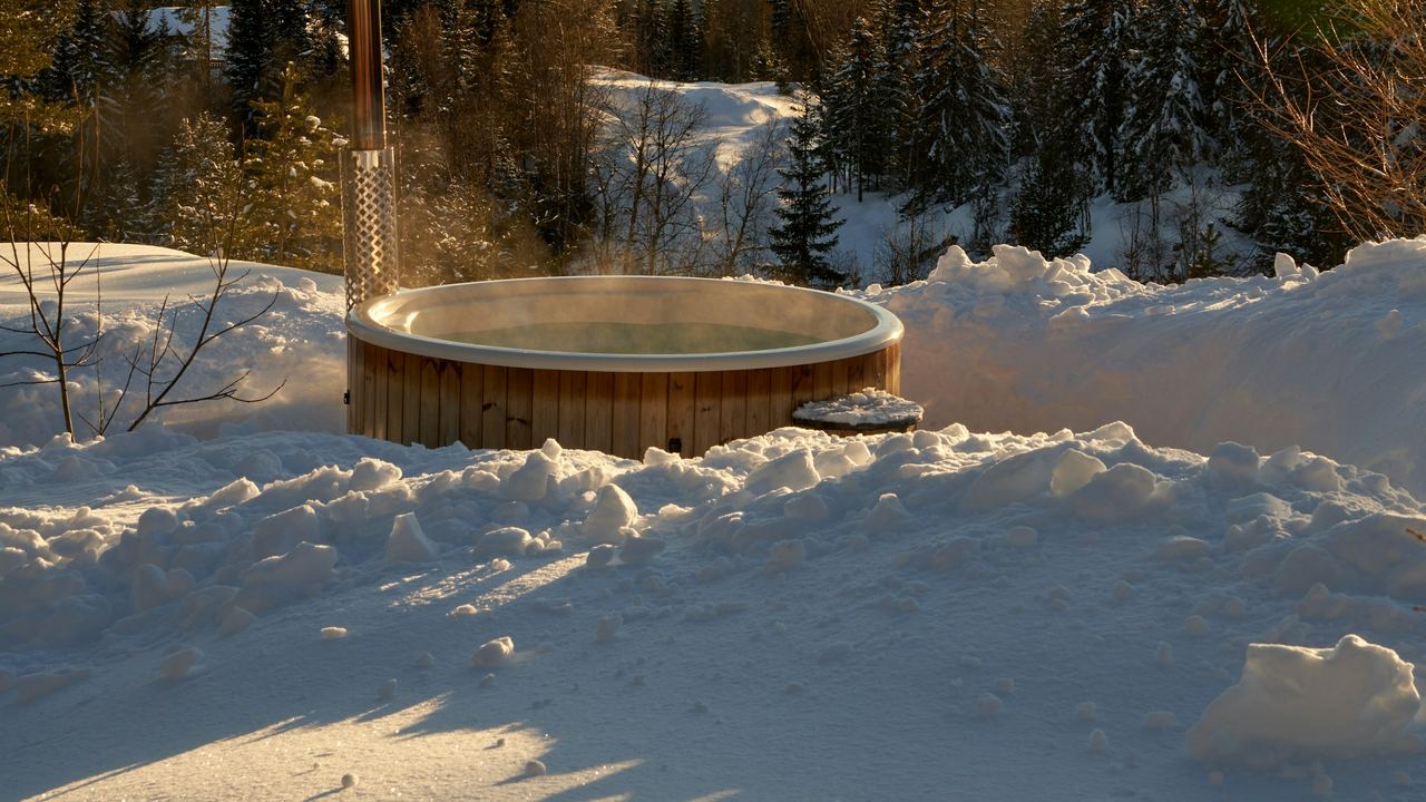 A steaming wood-fired hot tub in the snow, with forested mountains beyond