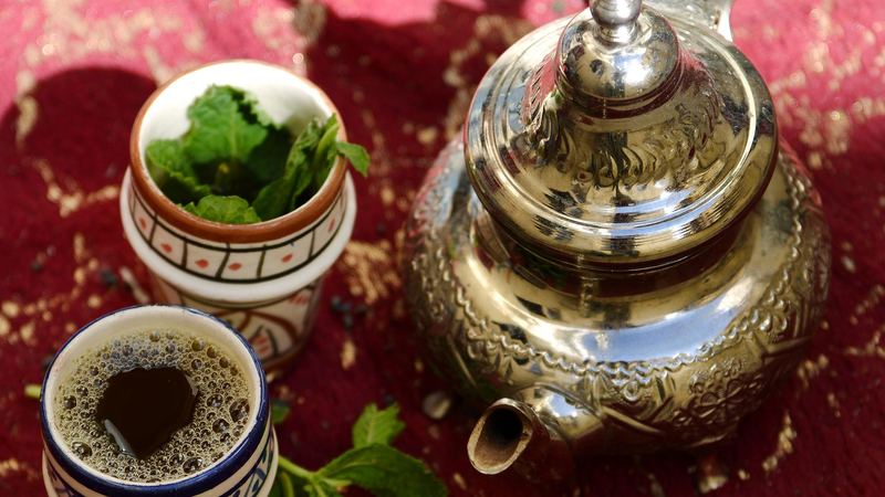 An engraved Moroccan teapot beside a pot of fresh mint and a cup of tea on a red cloth