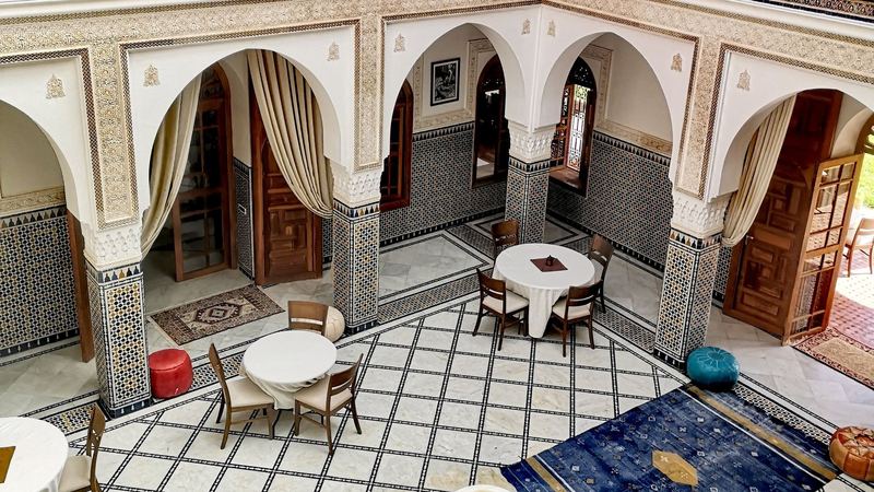 A tiled riad courtyard with carved arches, a marble fountain and tables set for tea
