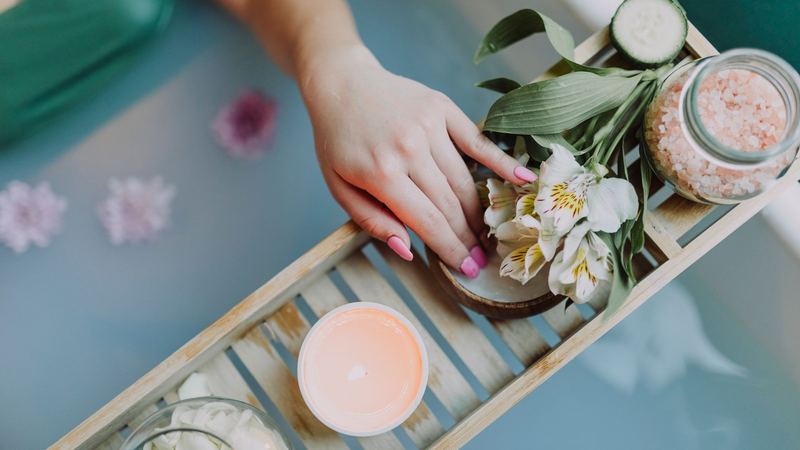 A hand resting on a bath tray of flowers, bath salts and a candle above a bath strewn with petals