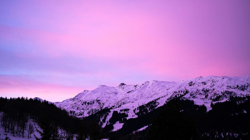 A crescent moon in a pink and violet sky above the snowy slopes and dark forests of the valley