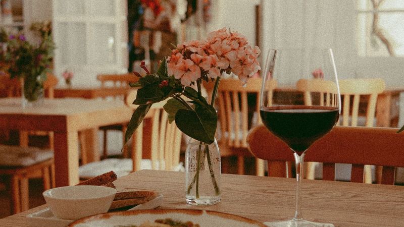 A glass of red wine, pink flowers in a vase and bread on a wooden table in a bright restaurant
