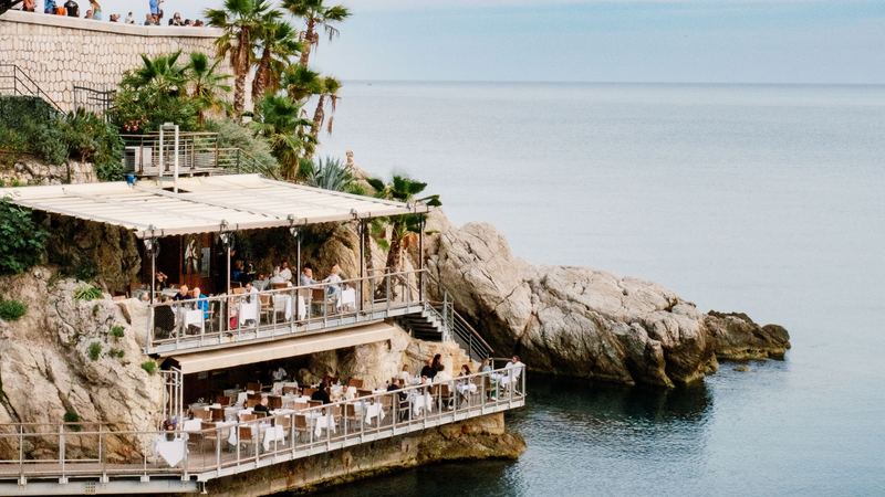 A restaurant on two terraces built into the rocks above a calm sea, palms and a stone wall above it