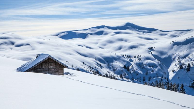 A wooden alpine hut half buried in snow on a sweeping white slope