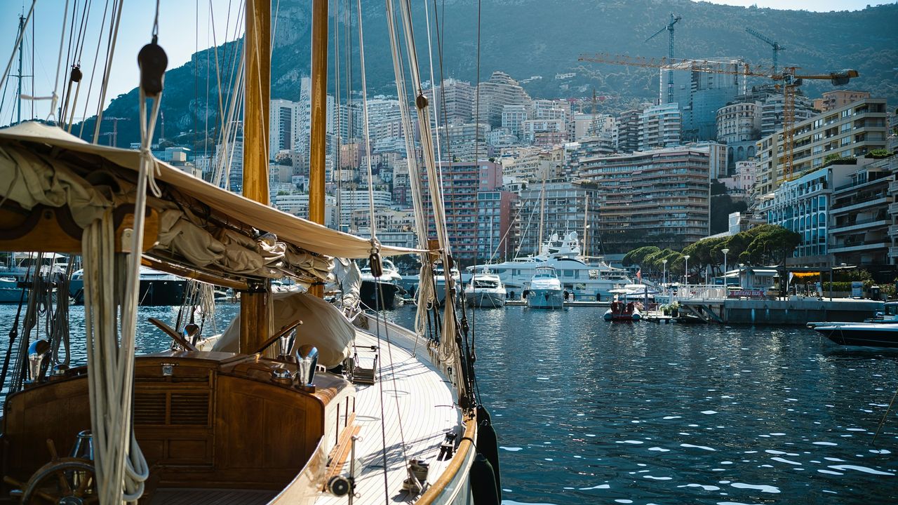 The varnished deck and wooden masts of a classic sailing yacht in Port Hercule, the towers of Monte-Carlo beyond