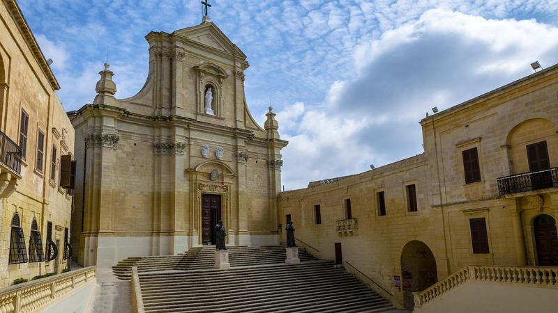 The Baroque front and wide stone steps of the Cathedral of the Assumption inside the Citadel of Gozo under a blue sky