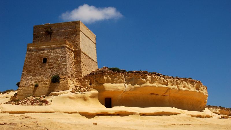 An old stone watchtower on golden sandstone cliffs on the coast of Gozo under a deep blue sky