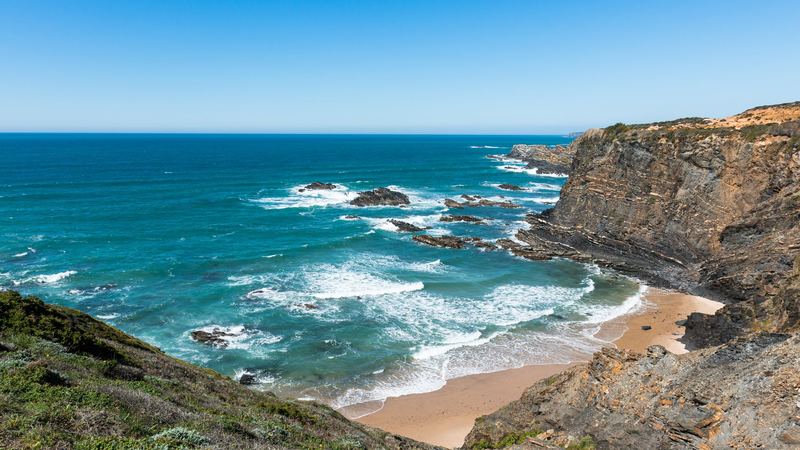 A sandy cove below layered cliffs, with turquoise Atlantic waves breaking on the rocks