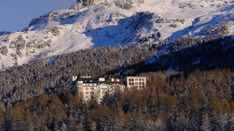 A grand hotel flying the Swiss flag among snowy larch forest below a mountain in winter sun