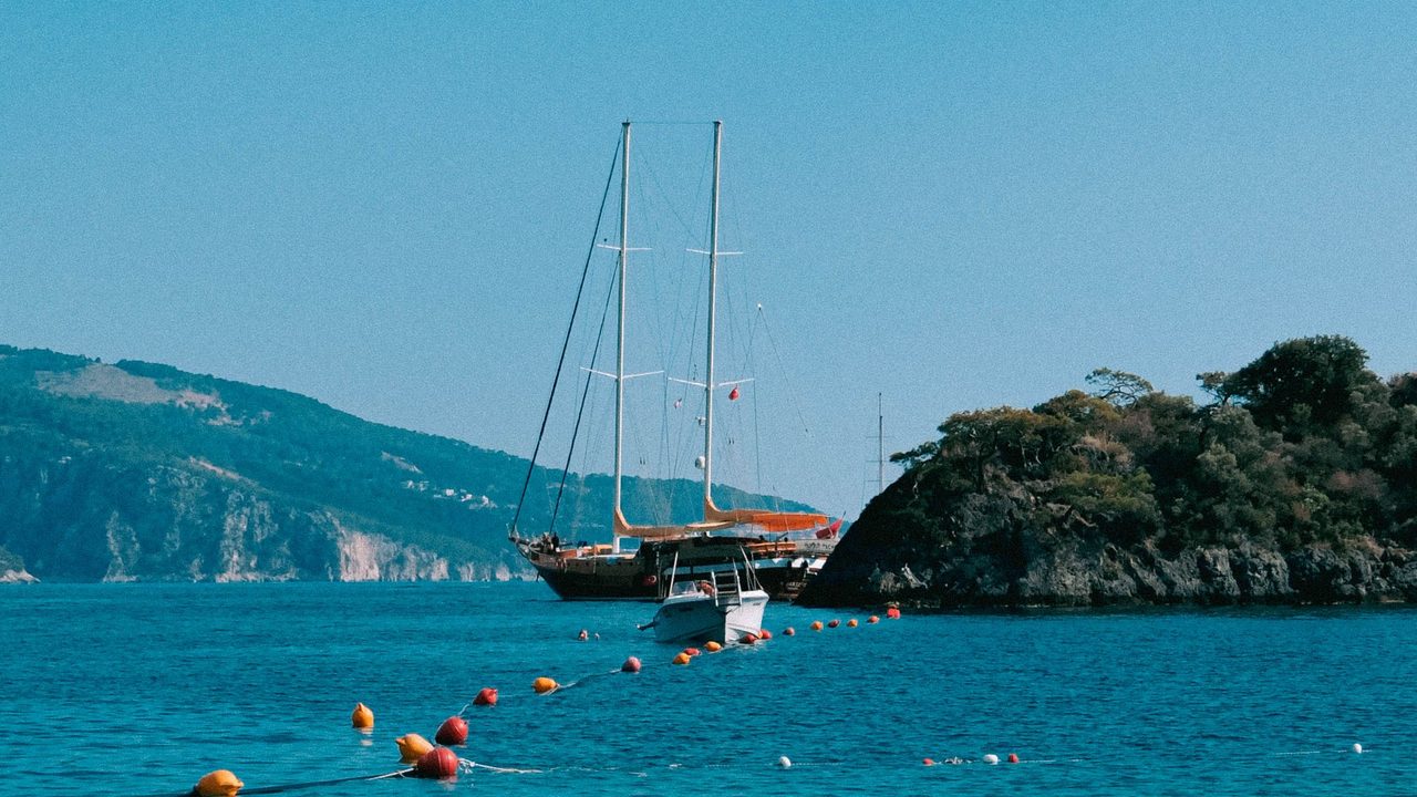 A two-masted wooden gulet at anchor in a clear turquoise bay below wooded hills